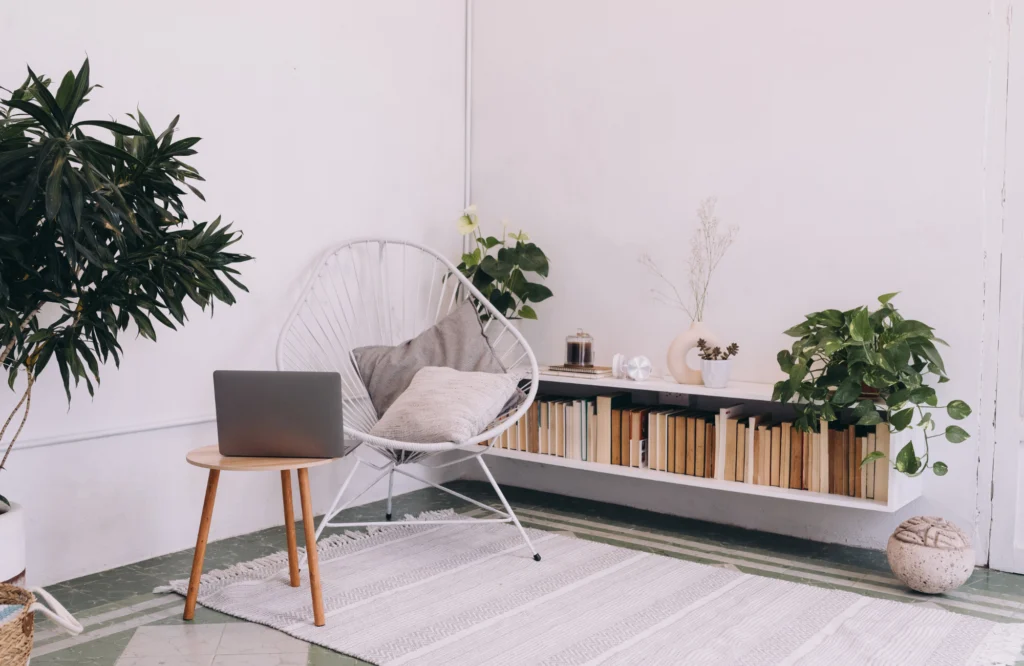 Modern seating area featuring a white chair, laptop on a wooden table, and a low built-in bookshelf with neatly arranged books, complemented by indoor plants and a light rug, illustrating effective bookshelf styling and organization tips.