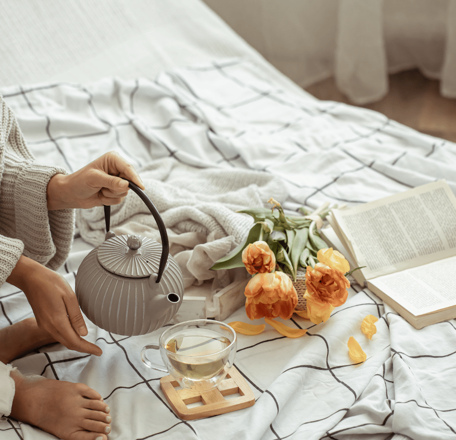 Cozy scene featuring a person pouring tea from a gray kettle into a glass cup, surrounded by yellow tulips, a blanket, and an open book on a bed, reflecting themes of self-care and kindness.