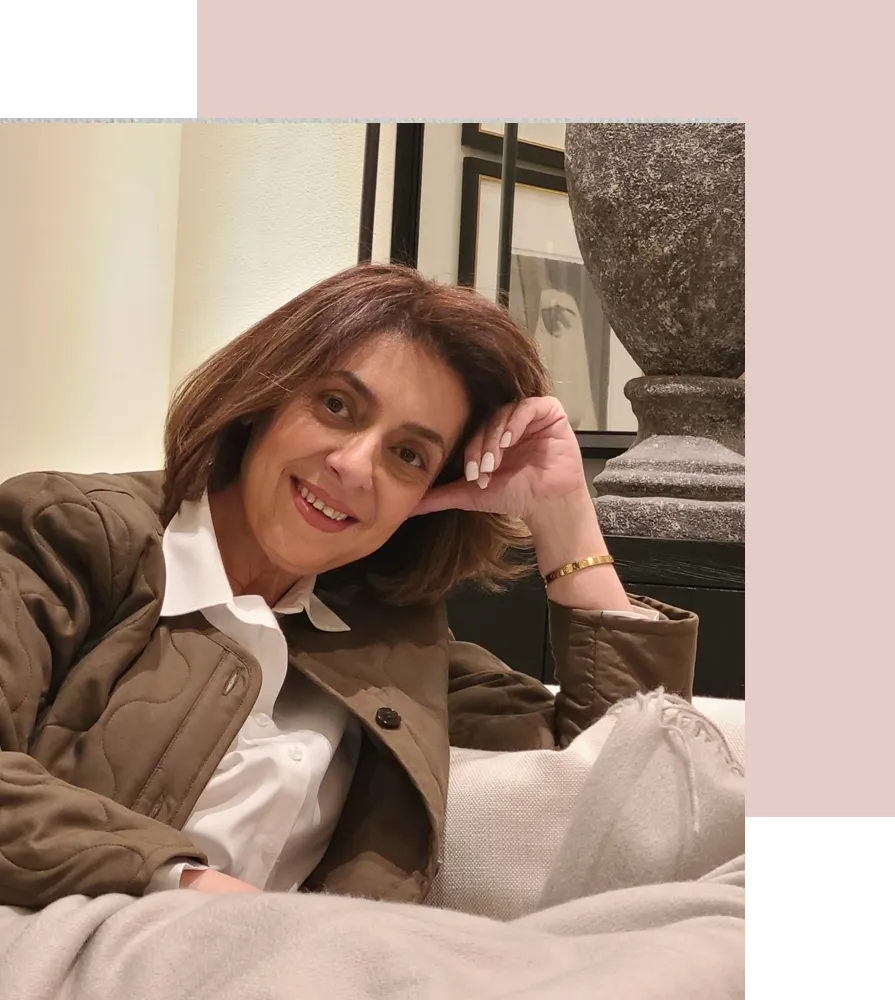 Woman smiling and relaxing on a sofa, wearing a stylish jacket and white shirt, representing a holistic and personalized approach to life coaching and productivity enhancement.