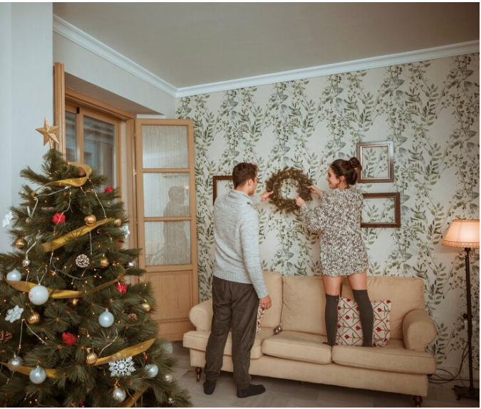 Couple decorating living room for holidays, hanging wreath on wall, Christmas tree adorned with ornaments in foreground, cozy atmosphere.
