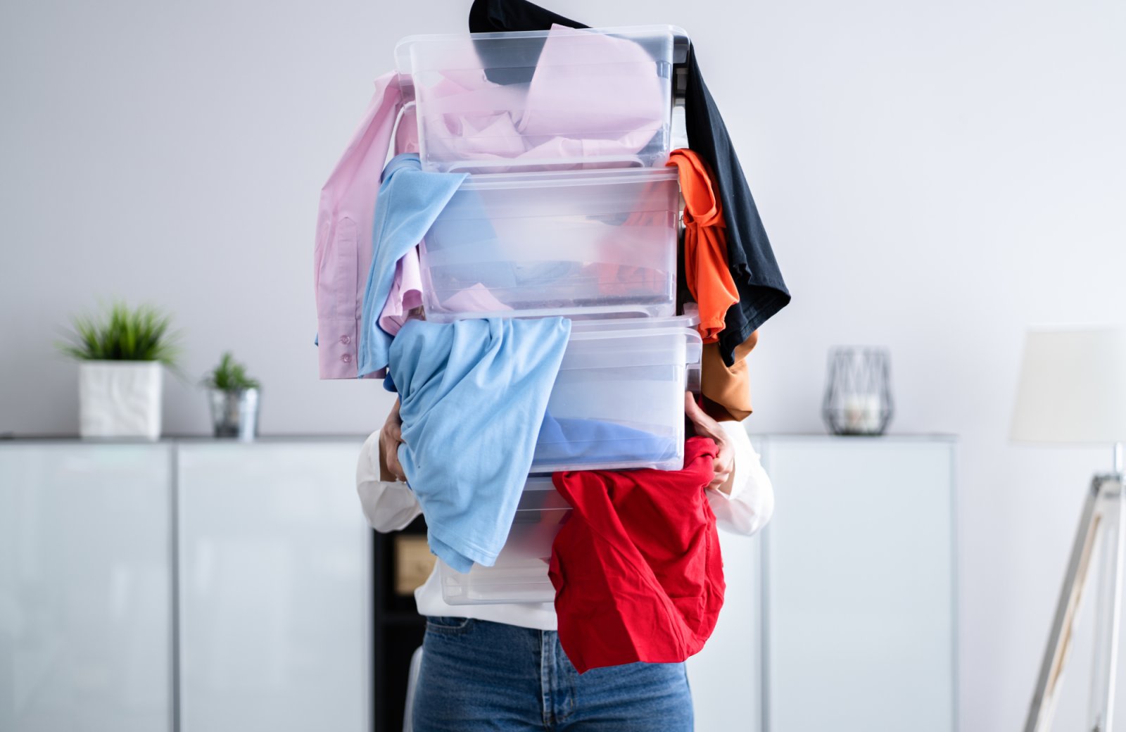Woman holding stacked bins filled with colorful clothing, illustrating the decluttering process before listing a home for sale, emphasizing organization and visual appeal.